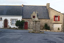 Photographie de trois maisons basses couvertes d’ardoise derrière une croix de granit.