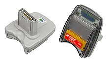 An image of the front and back of the Transfer Pak accessory. The front shell is made of gray plastic with a Nintendo 64 logo printed on it, and features a long protrusion with a rectangular opening at the end; a small set of electrical contacts is visible within the opening, which acts as the connection point when the device is inserted into the Nintendo 64 controller. The back shell is made of transparent plastic and features a thin opening at the top, with a large gap inside the shell shaped to the dimensions of a Game Boy cartridge; at the bottom of the gap is the Game Boy cartridge reader. A thin red sticker on the back displays the device's name, model number, and assorted legal indica.