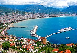 A colorful city with red roofs rising out from a curving harbor with blue water and cruise ship docked by a long pier.