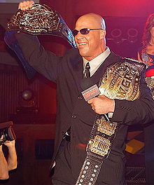 *** white male wearing a black suit and sunglasses holding two black belted championship belts and a microphone.